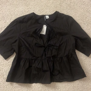Svart blus med knyt från H&M - Svart blus från H&M med knytning framtill och lätt volang nertill. Modellen har korta ärmar och en rundad halsringning. Perfekt att styla med jeans eller kjol för en trendig look. Oanvänd med prislapp!