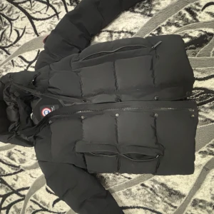 Svart dunjacka från Canada Goose - Säljer en svart dunjacka från Canada Goose med klassisk patch på ärmen. Jackan har huva, stora fickor framtill och dragkedja. Perfekt för kalla dagar med sin tjocka, quiltade design och stilrena look.