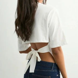 Vit topp med knytband  - Säljer en vit croppad topp med korta ärmar och snygg öppen rygg som knyts med breda band. Toppen har en enkel och clean look framtill och är tillverkad i mjuk bomull. Perfekt för dig som vill ha en trendig och luftig stil.