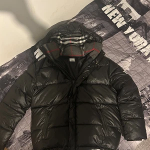 Svart pufferjacka Burberry London England - Svart pufferjacka från Burberry London England med huva och dragkedja. Insidan av kragen har ett snyggt rutigt mönster i grått, vitt och rött. Jackan har två fickor med dragkedja och en klassisk Burberry-logga på ärmen. Perfekt för kalla dagar.