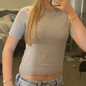 Grå croppad topp från 157 - Säljer en ljusgrå croppad topp från 157 med korta ärmar och rund halsringning. Toppen är stickad i mjukt material och har ribbade kanter vid ärmslut och nederkant. Perfekt till jeans eller kjol för en enkel och clean look.
