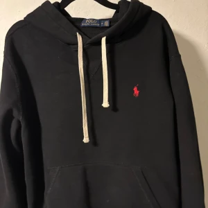Svart hoodie från Polo Ralph Lauren - Svart hoodie från Polo Ralph Lauren med vit snörning och klassisk röd logga broderad på bröstet. Tröjan har huva, känguruficka och är tillverkad i mjuk bomull. Perfekt för en chill och stilren look.