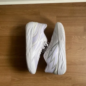 Vita sneakers från Puma med mönster - Säljer ett par sprillans nya, vita LaMelo från Puma med stilrena grå detaljer och coolt grafiskt mönster på sidorna. Skorna har snörning, platt sula och är tillverkade i syntet och textil. Perfekta för dig som vill ha skor till basket, paddel och annan träning. 🤍🤍