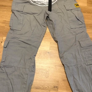 Grå cargopants från Corteiz i bomull - Säljer ett par grå cargopants från Corteiz i 100% bomull. Byxorna har flera stora fickor på sidorna och bak, samt en gul Corteiz-patch. Perfekta för en streetwear-look och riktigt sköna att bära.