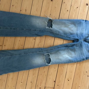 Blå bootcut jeans från Nelly, strl 36 - Bootcut jeans i ljusblå denim från Nelly med slitna hål på båda benen. Klassiska bakfickor med lock och knapp, samt snyggt tvättad finish. Perfekta för en avslappnad och trendig look.