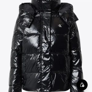 Svart pufferjacka från Calvin Klein - Säljer en glansig svart pufferjacka från Calvin Klein med hög krage och diskret CK-logo på bröstet. Jackan har quiltad design, dragkedja framtill och är perfekt för kalla dagar. Snygg och modern look som håller dig varm hela vintern.