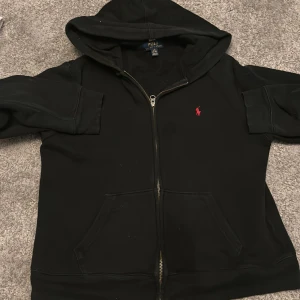 Svart hoodie från Polo Ralph Lauren - Svart hoodie från Polo Ralph Lauren med dragkedja och huva. Klassisk röd broderad logga på bröstet och stora fickor framtill. Perfekt för en chill och stilren look. Tillverkad i mjuk bomullsmix som känns skön mot huden.