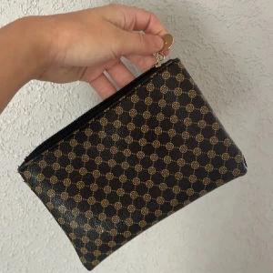 Svart och brun mönstrad plånbok - Snygg plånbok från shein i svart och brunt med klassiskt rutmönster och logga. Väskan har en praktisk dragkedja upptill och är tillverkad i skinn. Perfekt storlek för kort och småsaker, enkel att ta med överallt.