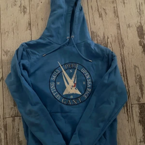 Blå hoodie från GANT med tryck - Säljer en blå hoodie från GANT med stort broderat tryck framtill med segelbåt och texten 'Setting Sails 1949 Open Sea Race'. Tröjan har huva med snörning och klassisk känguruficka. Perfekt för dig som gillar sportig och avslappnad stil.