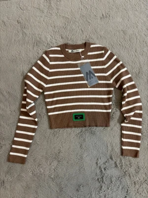 Brun och vit randig croppad tröja - Snygg croppad tröja från Met and March i brunt och vitt randigt mönster. Tröjan är ribbstickad, har långa ärmar och en rund halsringning. Detalj med grön patch nertill framtill. Perfekt för dig som gillar trendiga och bekväma plagg.