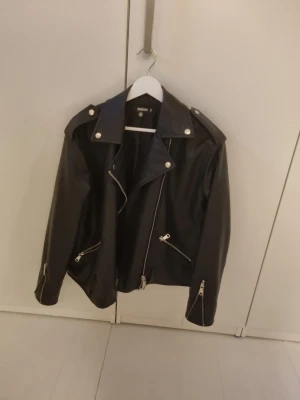 Svart skinnjacka från Missguided - Snygg svart skinnjacka från Missguided med coola silvriga dragkedjor och detaljer. Jackan har klassisk bikerstil med bred krage och axelklaffar. Perfekt för dig som vill ha en edgy look och gillar streetstyle.