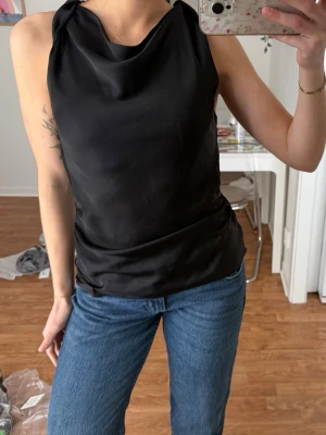 Svart satin topp från Zara - Snygg svart ärmlös topp från Zara i slätt, glansigt material med draperad halsringning. Toppen har en elegant och enkel design som passar perfekt till jeans eller kjol. Materialet känns lyxigt och har ett fint fall över kroppen. Strl S