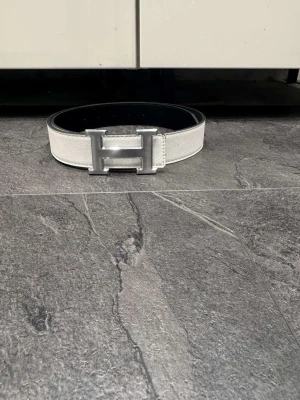Vit h-ermes bältd - Säljer ett stilrent skärp i vitt och svart med ett stort metalliskt H-spänne. Skärpet är gjort i präglat skinn och har en clean look som passar till många outfits. Perfekt accessoar för dig som gillar att sticka ut med detaljer.