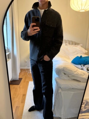 Mörkgrå overshirt i ullmix - Säljer en stilren mörkgrå overshirt med klassisk krage och knäppning framtill. Jackan har två bröstfickor med lock och knappar samt långa ärmar med knapp vid mudden. Perfekt lager-på-lager-plagg för höst och vår.