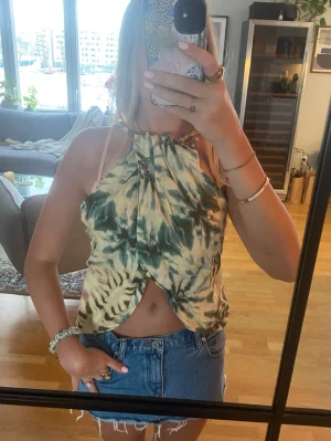 Grön och beige omlott linnetopp - Säljer en cool omlott linnetopp med knyt i halsen och öppet framtill. Toppen har ett batikmönster i grönt och beige med inslag av brunt. Materialet känns lätt och luftigt, perfekt för varma dagar. Passformen är loose och toppen är ärmlös med halterneck. På första bilden är den buren som det är tänkt men jag anser att det är snyggare bakåtvänt som visas på andra bilden 😻