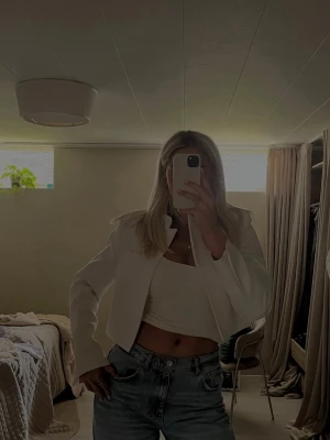 Vit croppad kavaj med ståkrage - Säljer en stilren vit croppad kavaj med snygg ståkrage och raka axlar. Jackan har en minimalistisk design utan synliga knappar eller dragkedja och är perfekt för dig som gillar en clean look. Passformen är normal och materialet känns strukturerat och lite tjockare.