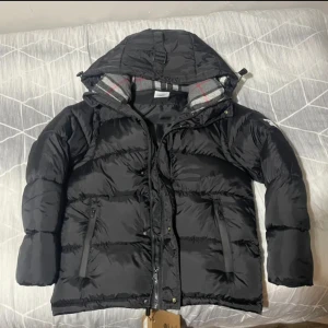 Svart pufferjacka från Burberry - Säljer en svart pufferjacka från Burberry med huva och dragkedja. Jackan har Burberry-logga på ärmen och klassiskt rutigt foder i halsen. Två fickor med dragkedja framtill och en schysst oversized look. Perfekt för kalla dagar och riktigt snygg till streetwear, jackan är helt ny.