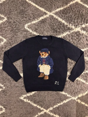 Polo Bear Ralph Lauren tröja  - Säljer en mörkblå stickad tröja från Polo Ralph Lauren med ikoniska Polo Bear-motivet framtill. Tröjan har rund hals, långa ärmar och RL-broderi vid nederkanten. Perfekt för dig som gillar klassisk preppy stil. Jag väger 70KG och är 180CM och den passar mig inte alls.