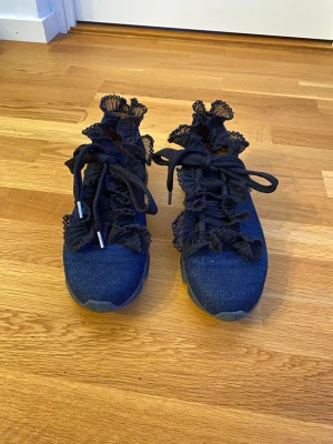 Blå sneakers med spets från Ganni - Unika sneakers från Ganni i mörkblå denim med svarta snören och dekorativ spets längs kanterna. Snygg platt sula och rund tå, perfekt för dig som vill sticka ut med coola detaljer. Insidan är i brunt skinn för extra komfort.