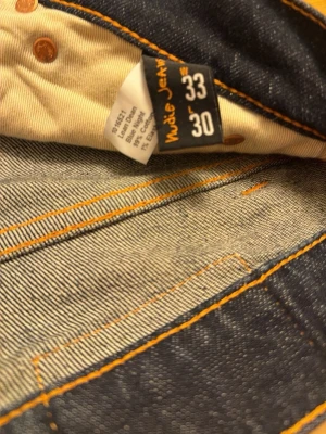 Mörkblå Nudie Jeans Lean Dean W33 L30 - Säljer ett par mörkblå Nudie Jeans Lean Dean med klassiska orange sömmar och snyggt bakficksmönster. Jeansen har normal passform och är tillverkade i stretchig denim av 99% bomull och 1% elastan. Perfekta för dig som gillar stilrena och bekväma jeans.