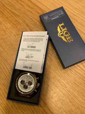 LIGE klocka med brun läderrem - Stilren klocka från LIGE med vit urtavla, guldfärgade detaljer och tre svarta subdials. Remmen är i brunt läder med krokopräglat mönster och klassiskt spänne. Perfekt accessoar för dig som vill ha en snygg och tidlös look. Den är helt ny skick säljer eftersom jag fick samma klocka flera gånger. Det går att förhandla priset.