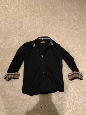Svart Burberry skjorta med rutiga detaljer - Säljer en svart skjorta från Burberry i storlek M. Skjortan har klassiska Burberry-rutiga detaljer på krage och manschetter, knappar framtill och lång ärm. Perfekt för dig som vill ha en stilren look med en twist.