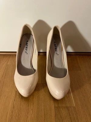 Beige pumps med platå från Kafeini - Säljer ett par stilrena pumps i beige lack från Kafeini. Skorna har hög klack och platåsula, rund tå och en snygg glansig finish. Perfekta för dig som vill ha en klassisk och elegant look. Storlek 37.