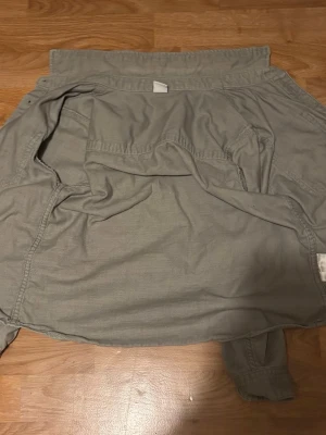 Grå grön overshirt med knappar - Säljer en grå overshirt i bomull med klassisk krage och stora bröstfickor. Jackan har långa ärmar med knapp vid ärmslut och knäppning längs hela framsidan. Perfekt för lager på lager och enkel att styla till olika outfits.