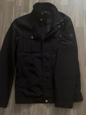 Jlindeberg field jacket  - Svart vindjacka från J.Lindeberg med coola detaljer som bröstfickor med knappar, dragkedja och emblem på ärmen. Jackan har hög krage och knappstängning framtill. Perfekt för dig som gillar stilren och sportig look. I perfekt skick! 