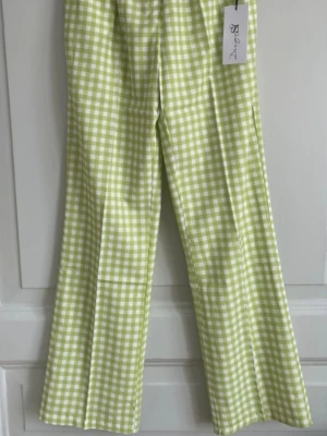 Grönrutiga byxor Design by Si M - Snygga byxor från Design by Si i storlek M. De har ett grönt och vitt rutigt mönster, rak passform och elastisk midja för extra komfort. Perfekta för dig som vill sticka ut med en fräsch och trendig look. Storlek 36/38(liten M)