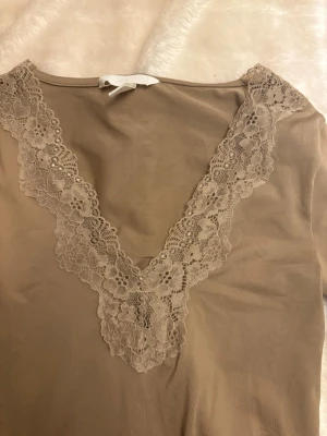Beige långärmad spetsbody - Säljer en beige body från H&M med lång ärm och djup v-ringning dekorerad med blommig spets. Materialet är mjukt och stretchigt, perfekt för att styla med jeans eller kjol. Spetsen ger en feminin touch och bodyn har en slim passform. xs - s