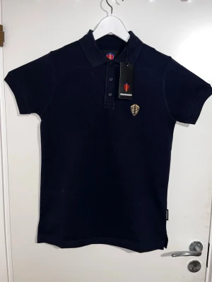 Koenigsegg piké - Säljer en marinblå pikétröja från Koenigsegg med guldfärgad broderad logga på bröstet och Koenigsegg-text broderad i gult på ryggen. Tröjan har klassisk krage, knappar framtill och är kortärmad. Materialet är piké i bomull som känns skönt mot huden. Nypris 950 mitt pris 399. Storlek XS men passar S och M