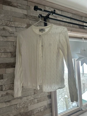 Vit kabelstickad kofta Hampton Republic - Vit kabelstickad kofta från Hampton Republic med rund halsringning och knappar framtill. Klassisk och enkel design med lång ärm och ribbade muddar. Perfekt att slänga över en topp för en clean look.