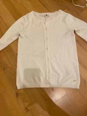 Vit kofta från Hampton Republic XS - Klassisk vit kofta från Hampton Republic i storlek XS. Koftan har rund hals, långa ärmar och knäppning framtill med små vita knappar. Tillverkad i mjuk bomull och har en liten guldfärgad logodetalj nedtill.