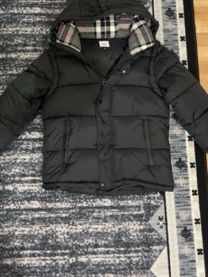 Svart pufferjacka från Burberry M - Säljer en svart pufferjacka från Burberry i storlek M. Jackan har klassiskt rutigt foder i huvan, dragkedja och knappar framtill samt två fickor med dragkedja. Materialet är polyester och jackan har en schysst, lite boxig passform.