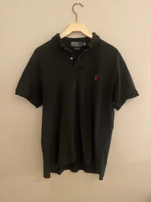 Svart piké från Polo Ralph Lauren - Klassisk svart pikétröja från Polo Ralph Lauren i slim fit. Tröjan har korta ärmar, två knappar vid halsen och den ikoniska röda loggan broderad på bröstet. Tillverkad i mjuk bomull som är skön att bära hela dagen.