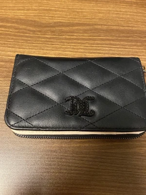 Juicy Couture svart quiltad plånbok - Juicy Couture plånbok i svart quiltad skinnimitation med logga framtill. Insidan är beige med flera kortfack, fönster för ID och ett myntfack med dragkedja och hjärtformad berlock. Fodret har Juicy Couture-tryck i svart. Perfekt för dig som gillar stilrena detaljer. Plånboken är inte använd, köpt för några år sedan men precis som ny! 
