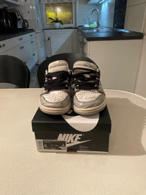 Nike Air Jordan 1 Low vit/svart - Nike Air Jordan 1 Low i vit och grå med svarta detaljer och swoosh. Klassisk siluett med svart snörning och perforerad tåbox. Skorna har en platt sula och är tillverkade i skinn och syntet. Perfekta för dig som gillar streetwear och sneakers med retro vibe.