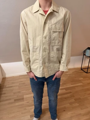 Beige overshirt från Peak Performance - Säljer en stilren beige overshirt från Peak Performance. Jackan har tre stora fickor framtill, klassisk krage och knäppning med knappar. Tillverkad i lätt bomullsmaterial, perfekt för lager på lager och enkel att matcha. Använd 1 gång. Nypris 1500
