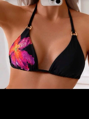 Svart bikiniöverdel med blomtryck - Säljer en svart bikiniöverdel med stort färgglatt blommönster i rosa, orange och rött på ena sidan. Överdelen har trekantsform och tunna band som knyts i nacken, dekorerade med guldfärgade ringar. Perfekt för stranden eller poolen!