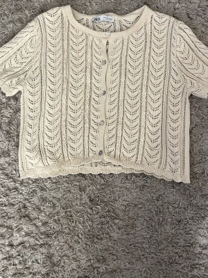 Beige stickad kofta från Zara - Säljer en beige, kortärmad stickad kofta från Zara med fina pärldetaljer och knappstängning framtill. Koftan har ett öppet, hålmönstrat stickat mönster och rund hals. Perfekt för vår och sommar när du vill ha en söt och luftig look.