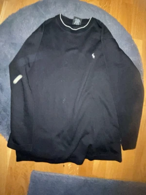 Svart långärmad tröja från Polo Ralph Lauren - Svart långärmad tröja från Polo Ralph Lauren med vit broderad logga på bröstet och en tunn vit kant vid halsringningen. Tröjan har en klassisk, normal passform och är tillverkad i mjuk bomull. Perfekt för chill dagar eller när du vill ha en clean look.