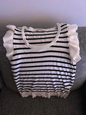 Randig topp med volangärm från Orsay - Snygg vit och svart randig topp från Orsay med volangdetaljer på axlarna. Toppen är ärmlös med rund halsringning och har en mjuk, bekväm känsla. Perfekt för en fräsch och trendig look.