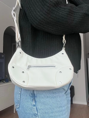 Vit axelremsväska med nitar, från H&M (nyskick) - Vit axelremsväska med dragkedja och platta nitar. Justerbar rem! Väskan har ett stort fack inuti samt ett litet på utsidan.  Storlek: 24x12cm  - Köper du innan kl 16:00 kan jag skicka samma dag!