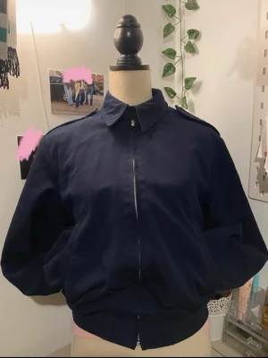 Mörkblå bomberjacka i ullmix - Säljer en mörkblå bomberjacka från RAF i storlek M. Jackan har klassisk krage, axelklaffar och dragkedja framtill. Tillverkad i en skön mix av polyester och ull, perfekt för dig som gillar en stilren och tidlös look.