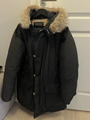 Svart parkajacka från Woolrich med pälskrage - Säljer en svart parkajacka från Woolrich med beige foder och stor huva med fluffig pälskant. Jackan har stora fickor framtill, knappar och dragkedja. Perfekt för kalla dagar och riktigt snygg till streetwear-stil.