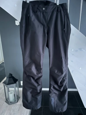 Svarta skidbyxor från Helly Hansen - Svarta skidbyxor från Helly Hansen i storlek XS. Byxorna har dragkedjor vid fickorna och knappstängning i midjan. Materialet är slitstarkt och vattenavvisande syntet, perfekt för kalla dagar. Diskret HH-logga på benet och klassisk rak passform.