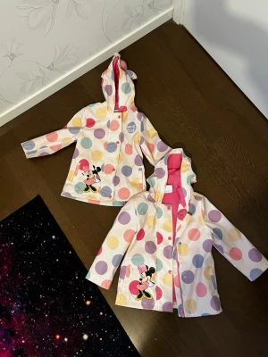 Minnie Mouse regnjackor - Supergullig regnjacka från Disney x Primark med färgglada stora prickar, Minnie Mouse-tryck och rosa innerfoder. Huvan har söta öron och en rosa rosett. Jackan stängs med knappar och har två fickor framtill. Perfekt för regniga dagar!