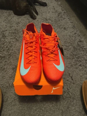 Nike fotbollsskor Vapor 16 PRO FG - Säljer ett par riktigt snygga Nike fotbollsskor i en skarp orange färg med ljusblå swoosh och rosa detaljer. Skorna har snörning, plös i samma färg och en sula med dobbar i rosa och blått. Ovandelen är i lätt syntetmaterial med texten FLYKNIT ACC på sidan.