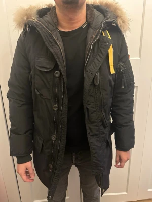 Svart Parajumpers parkas med pälskrage - Säljer en svart Parajumpers parkas med ikonisk gul rem, patch på ärmen och stor huva med pälskant. Jackan har flera fickor, dragkedja och knappar framtill samt detaljer som gör den riktigt snygg och funktionell för kalla dagar. Använd fåtal gånger